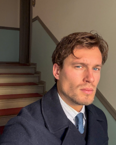 Man in a dark coat and tie standing in a room with a staircase.