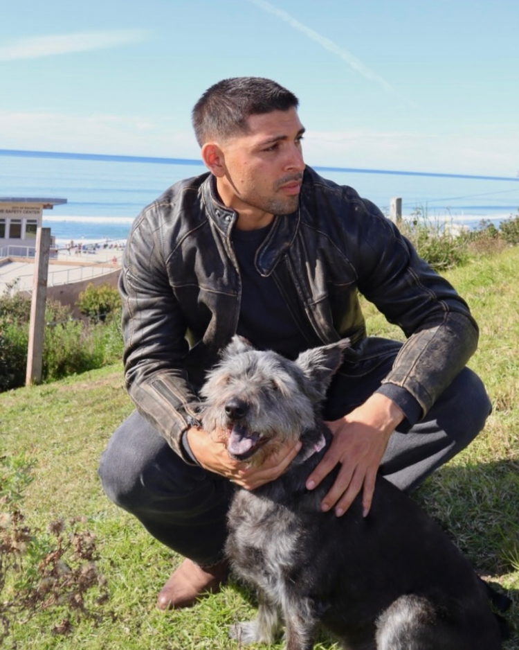 Man sitting on grass with a dog, both looking at the camera, with a coastal background.